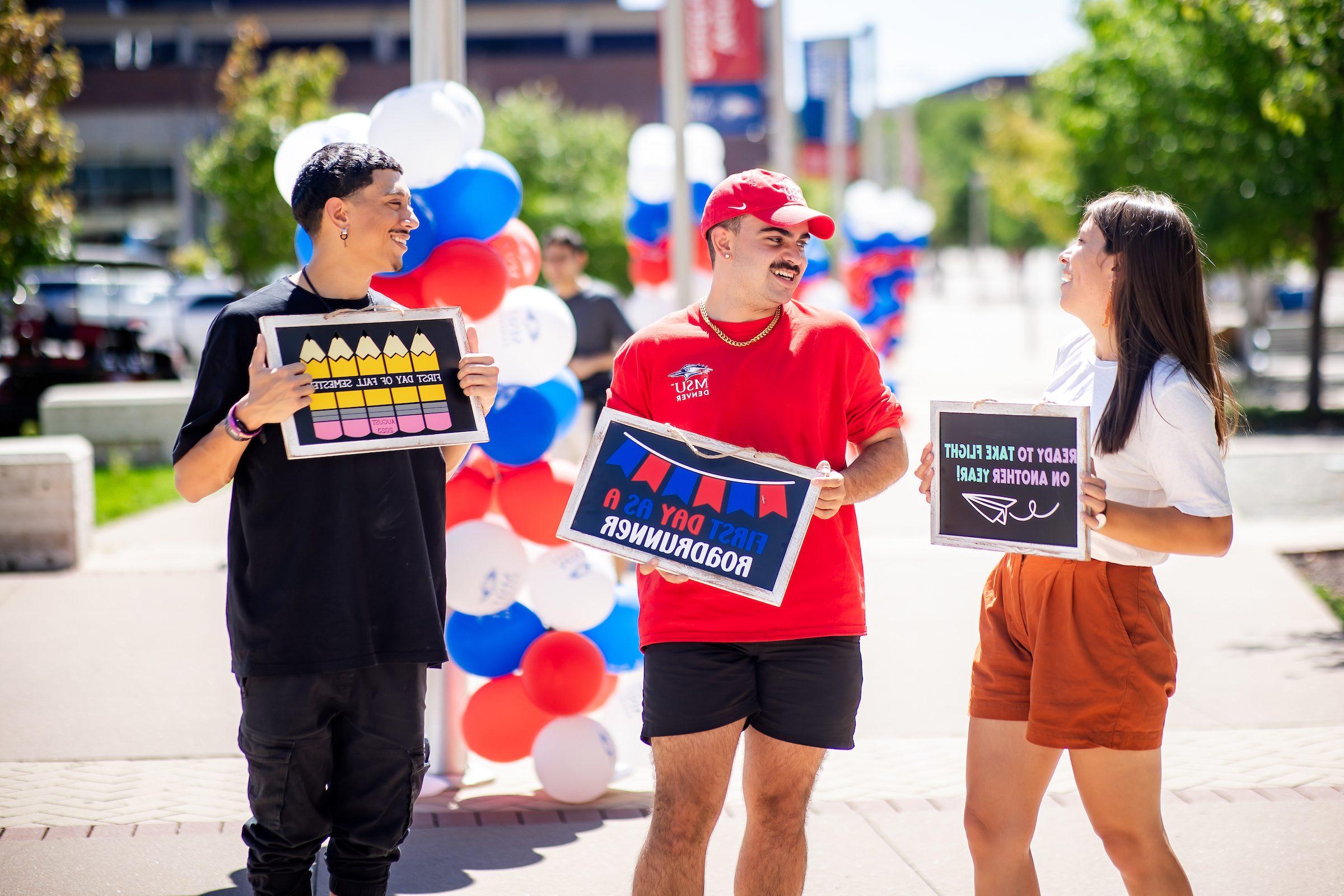 Three students together with signs welcoming students, smiling at each other during the first week of classes