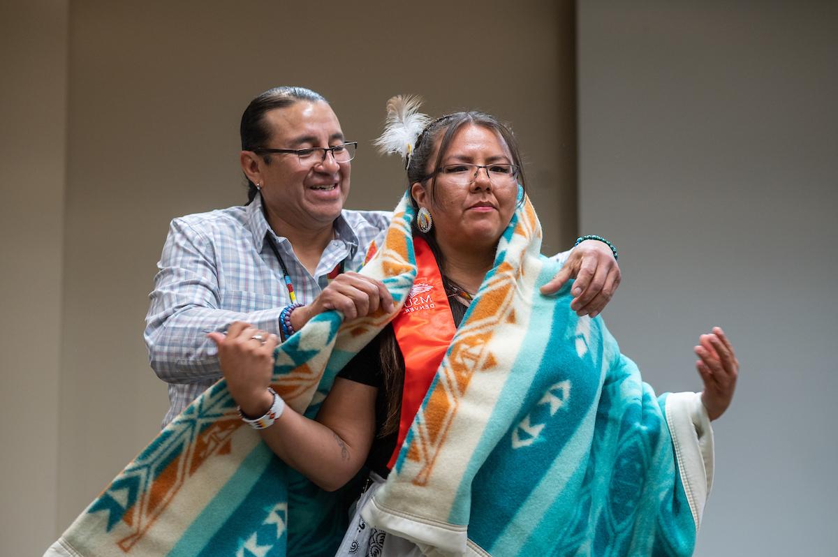 An older person wrapping a ceremonial Native-Indigenous blanket around a graduating MSU Denver student